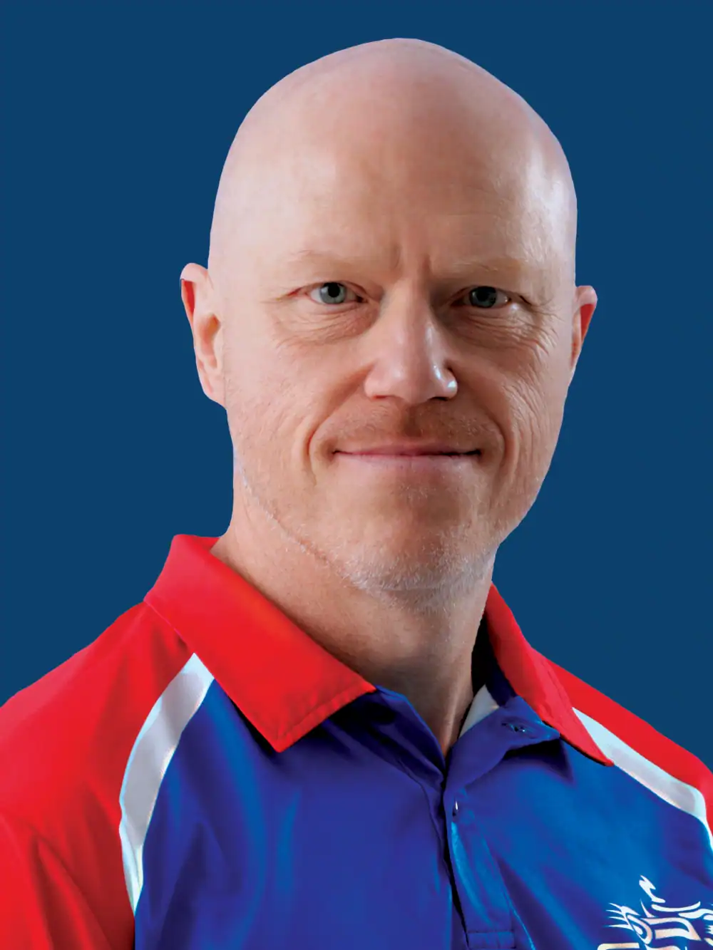 A bald man with a trimmed beard wearing a red, blue, and white collared sports shirt poses against a solid blue background.