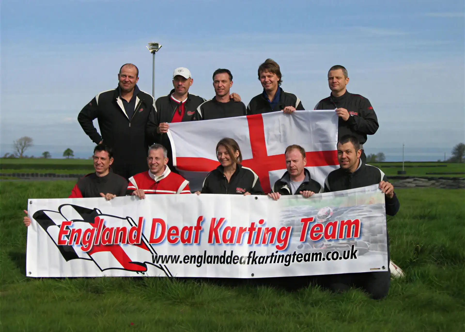A group of nine people in racing suits pose outdoors holding an England flag and a banner reading "England Deaf Karting Team".