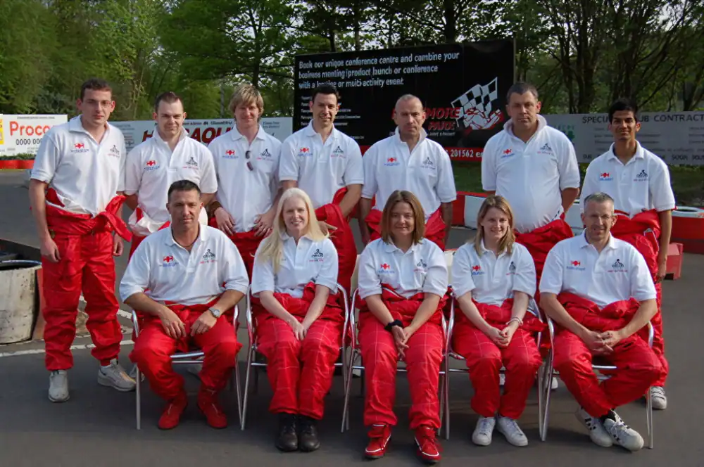 Twelve people in matching white polo shirts and red trousers pose outdoors in two rows; some are standing and others are seated on chairs. Trees and a black sign are visible in the background.