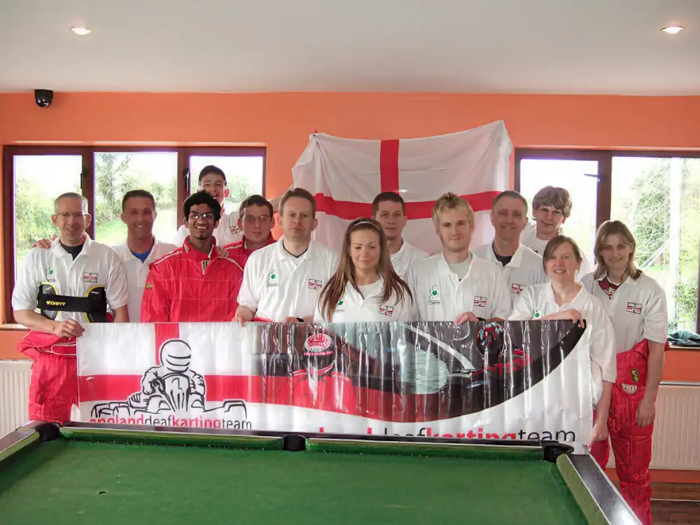 A group of people in matching England karting team shirts pose indoors, holding a team banner in front of a St George's flag.