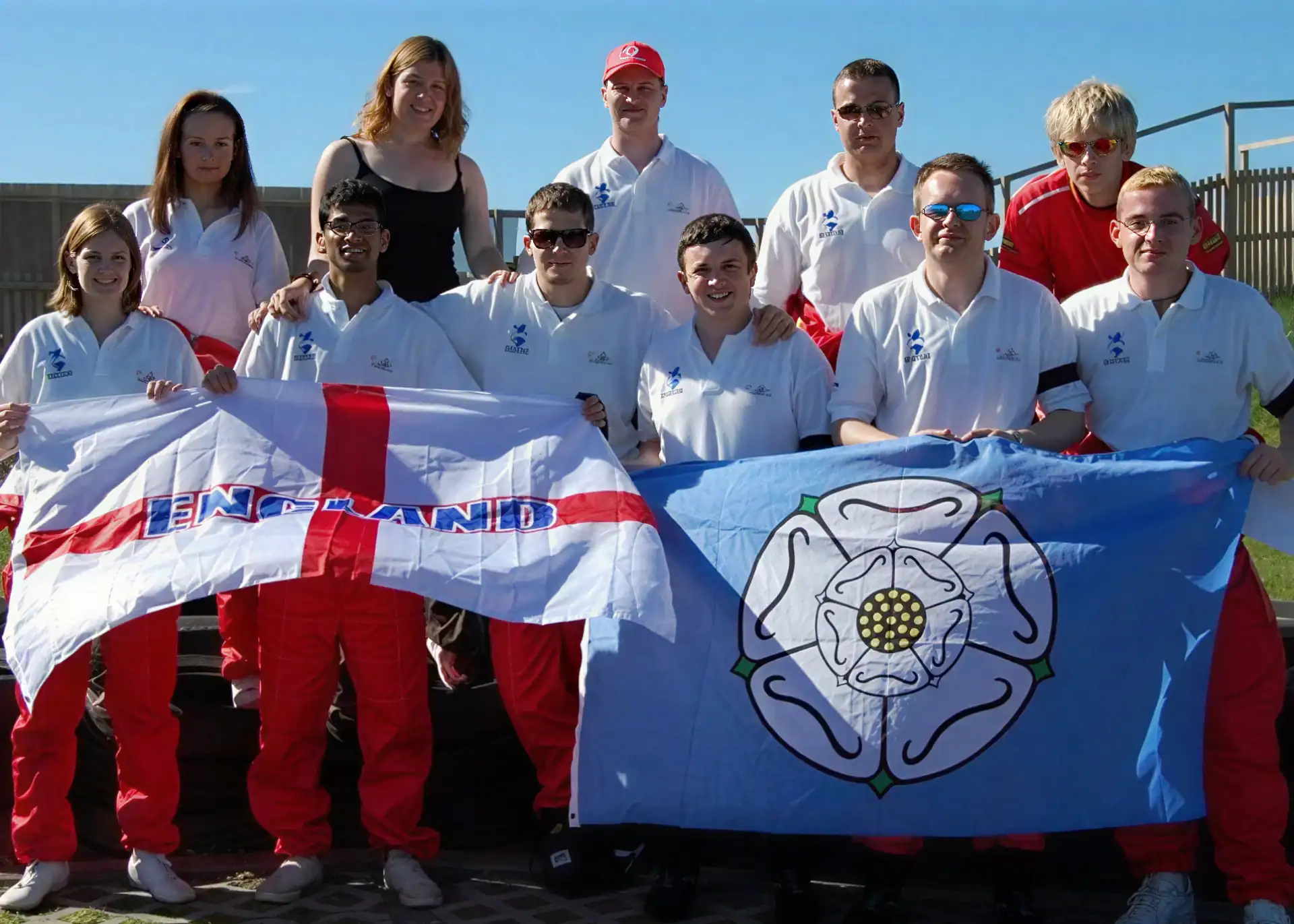 A group of people in white shirts and red trousers pose outdoors, holding an England flag and a blue flag with a white rose emblem.