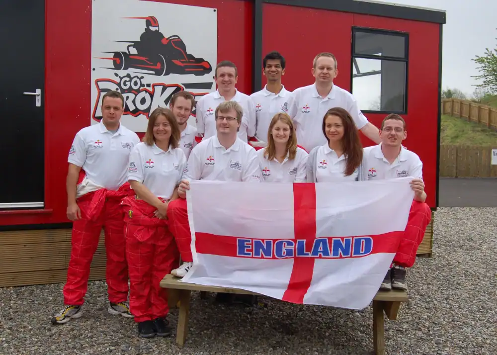 A group of ten people in matching white shirts and red trousers pose outside a building, holding an England flag with the word "ENGLAND" printed across it.