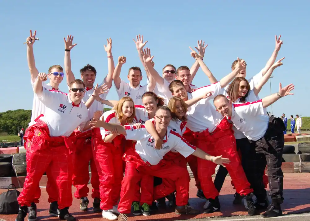A group of people in matching white shirts and red trousers pose enthusiastically outdoors, raising their arms and smiling on a sunny day.