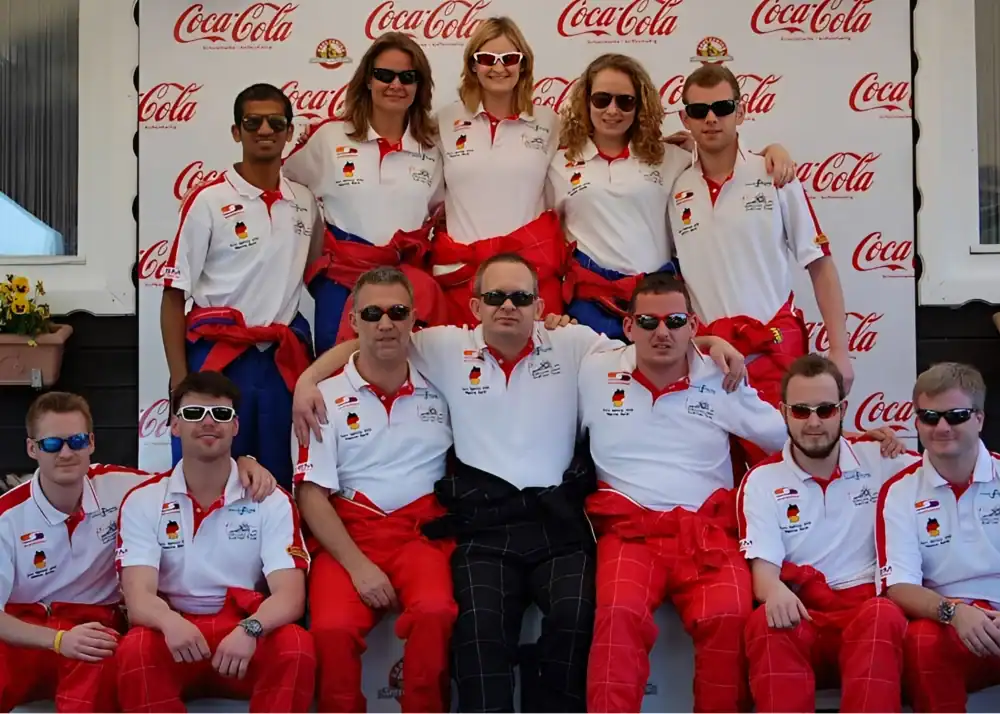 A group of eleven people in matching white shirts and red trousers pose for a team photo in front of a Coca-Cola branded backdrop.