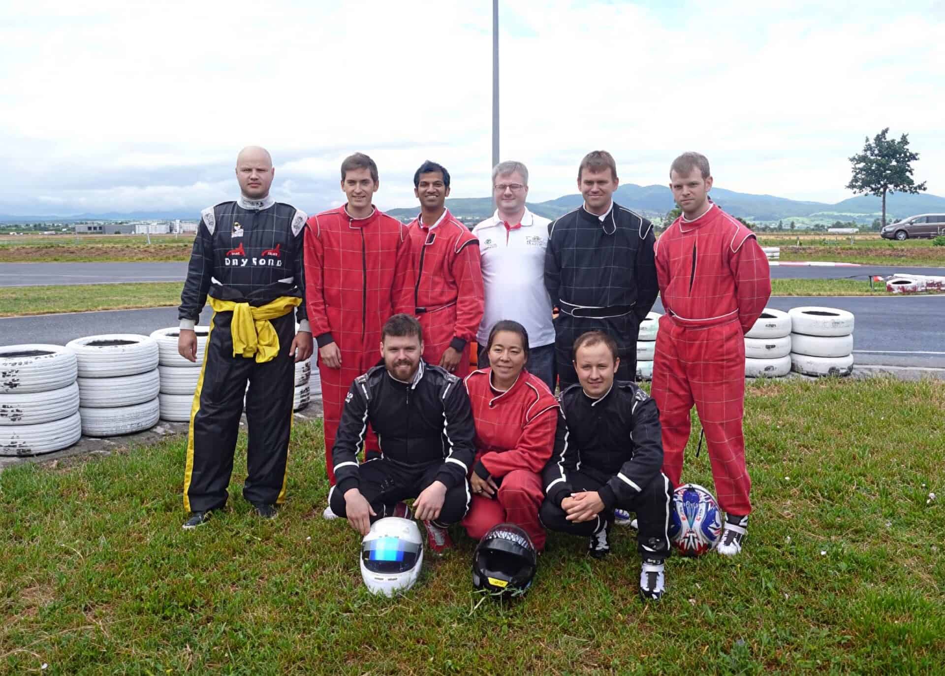 Nine people in racing suits pose on a grass area by a go-kart track with stacks of white tyres in the background; three hold helmets.