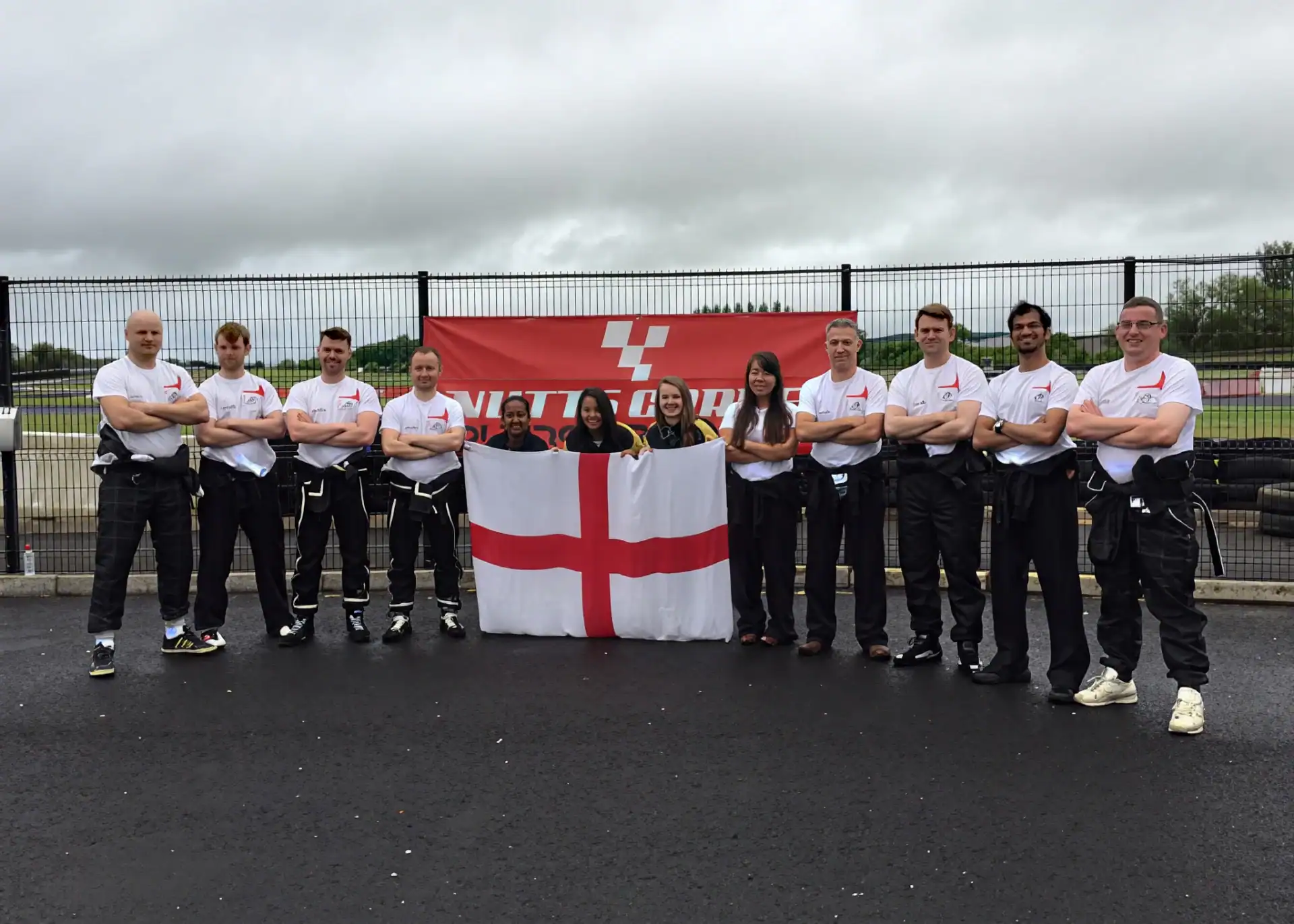 A group of twelve people in racing uniforms stand in front of a red banner and hold an England flag at an outdoor track.
