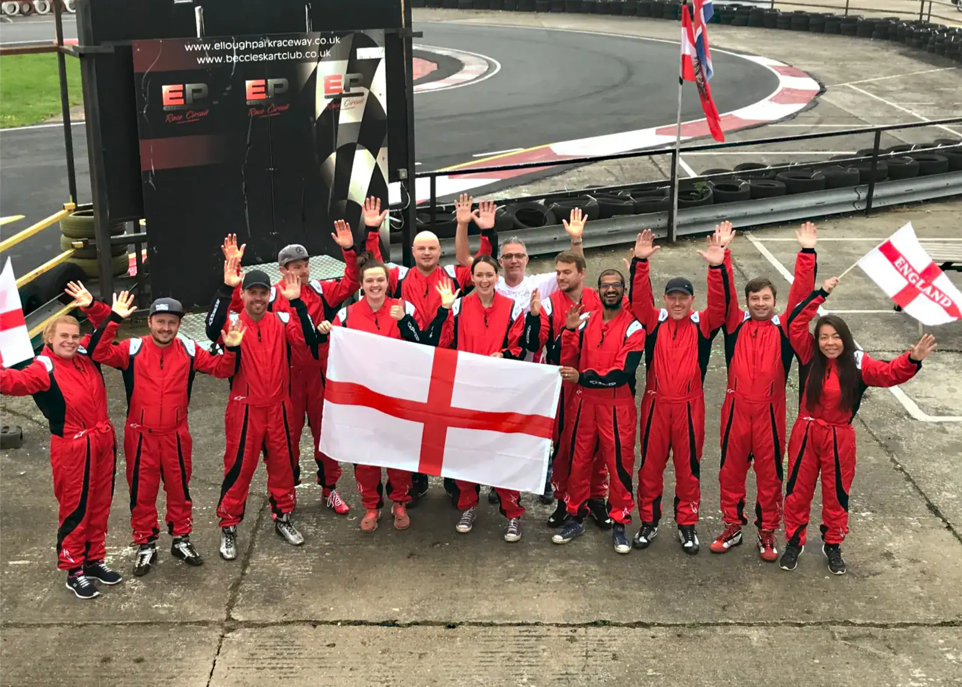 A group of people in red racing suits pose with an England flag and small banners on a go-kart track, raising their arms and smiling at the camera.