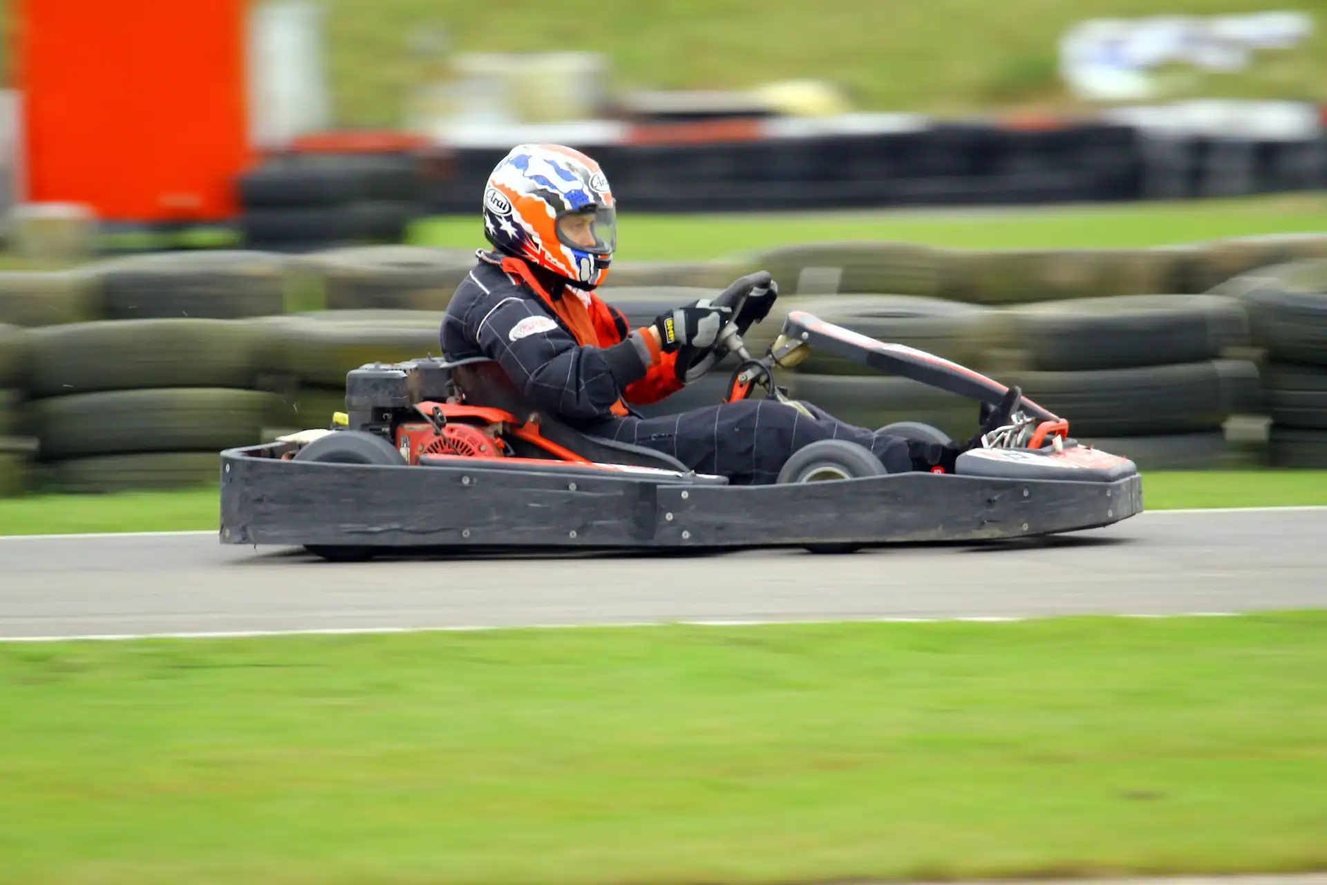 A person wearing a helmet and racing suit drives a go-kart on an outdoor track, with tyres and grass visible in the background.
