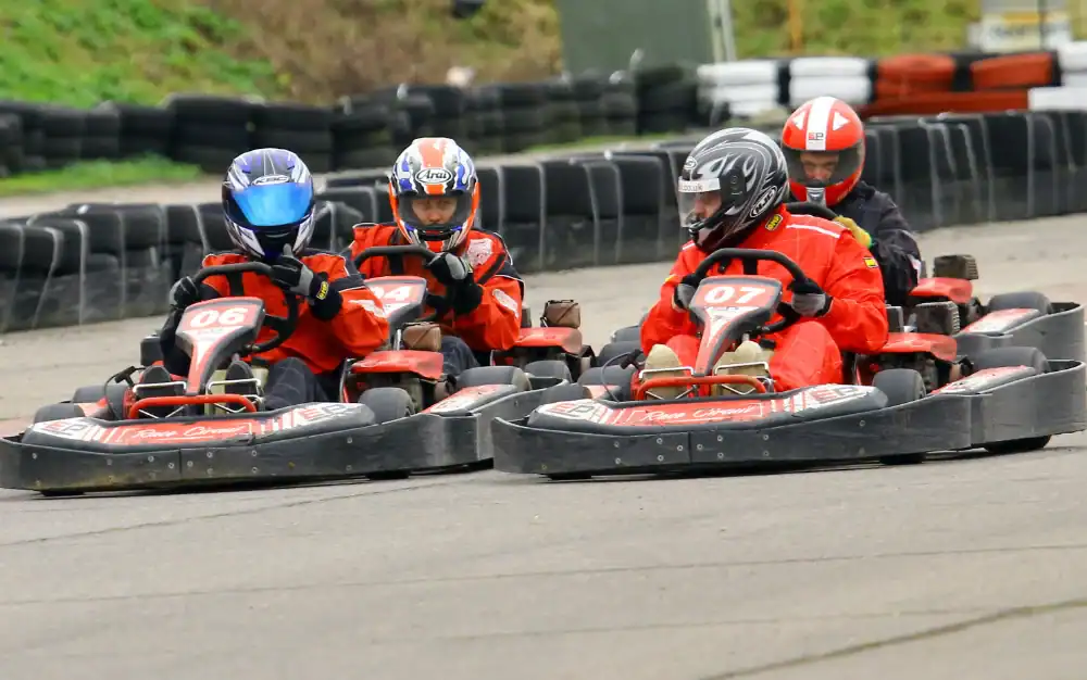 Four people wearing helmets and numbered racing suits drive go-karts closely together on an outdoor track with tyre barriers in the background.