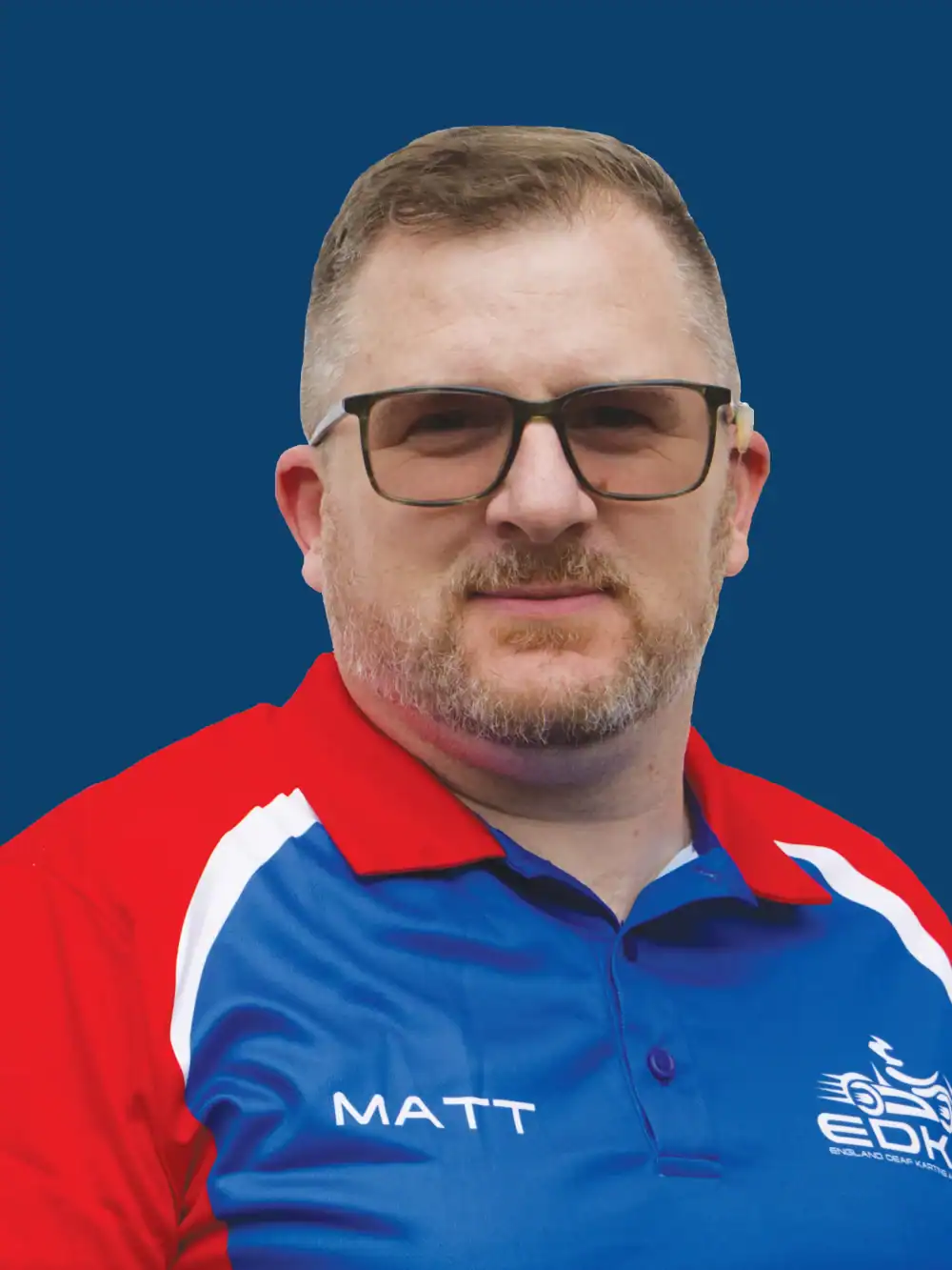 A man with short hair and glasses wearing a red, blue, and white collared shirt with "MATT" and a logo on it, standing in front of a solid blue background.