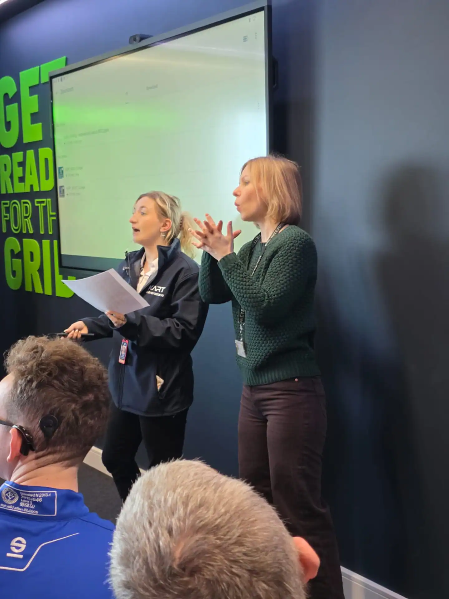 Two women stand at the front of a room, one speaking whilst holding papers, the other using sign language; a group of people listen. A large screen and wall text are visible behind them.