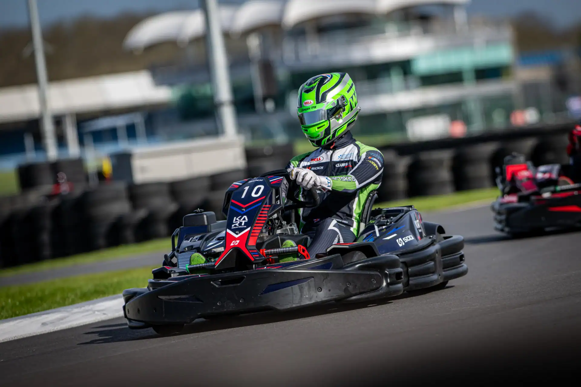 A person wearing a green helmet and racing suit drives a black go-kart with the number 10 on a racetrack, with another go-kart nearby.
