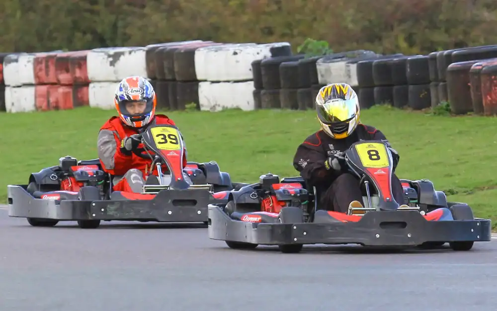 Two people wearing helmets drive go-karts numbered 39 and 8 on an outdoor track with tyre barriers in the background.