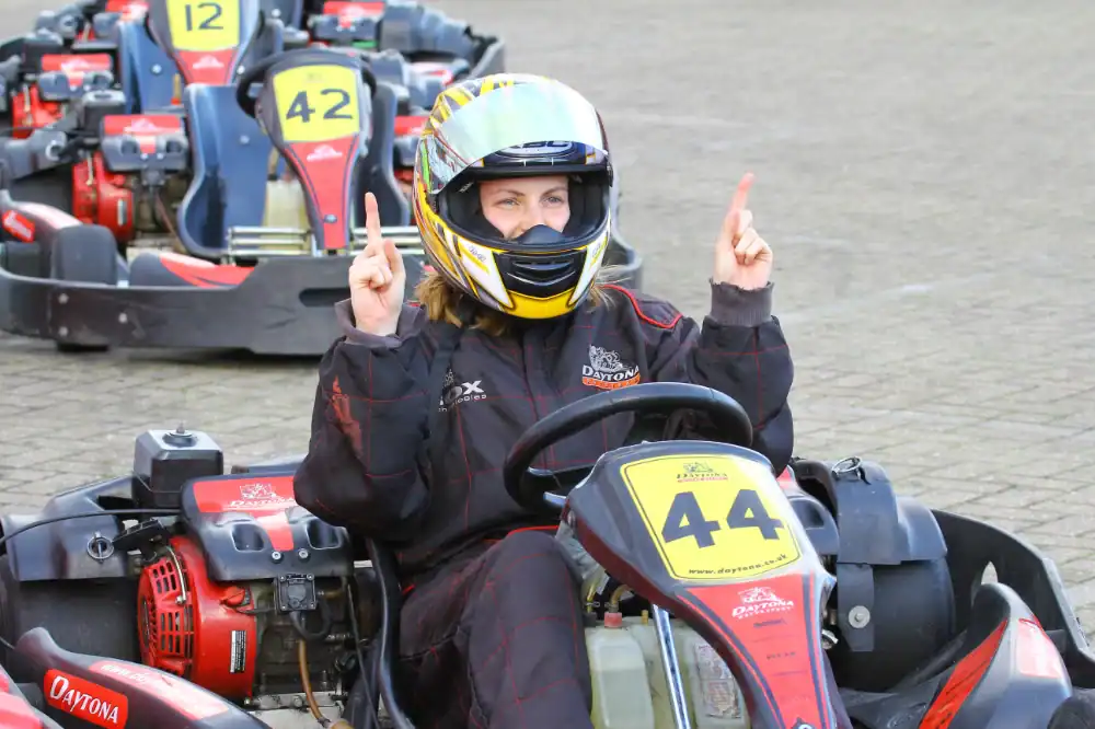 A person in a racing suit and helmet sits in a go-kart labelled 44, raising both index fingers, with other go-karts visible in the background.