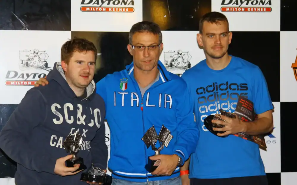 Three men stand in front of a Daytona Milton Keynes backdrop, each holding trophies and awards, posing for a photo after a competition or race event.