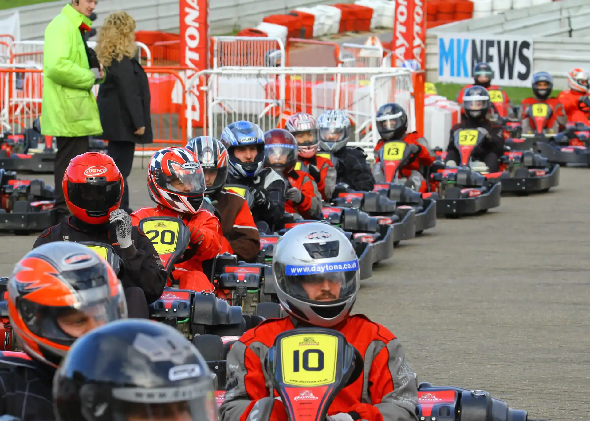 People wearing helmets and racing suits sit in numbered go-karts lined up on an outdoor track, preparing for a race. Barriers and signs are visible in the background.
