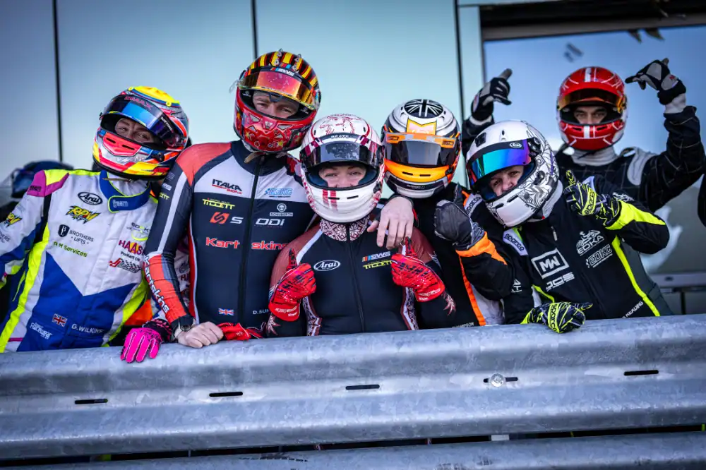Six go-kart racers in colourful racing suits and helmets pose together behind a metal barrier, with a building in the background.