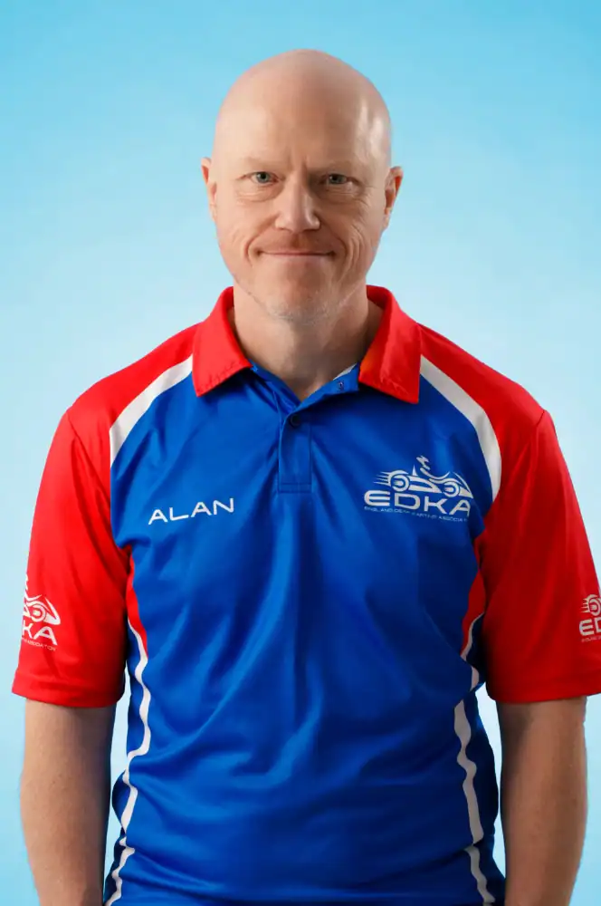 A bald man wearing a blue, red, and white polo shirt with "ALAN" and an "EDKA" logo stands against a light blue background, facing the camera and smiling slightly.