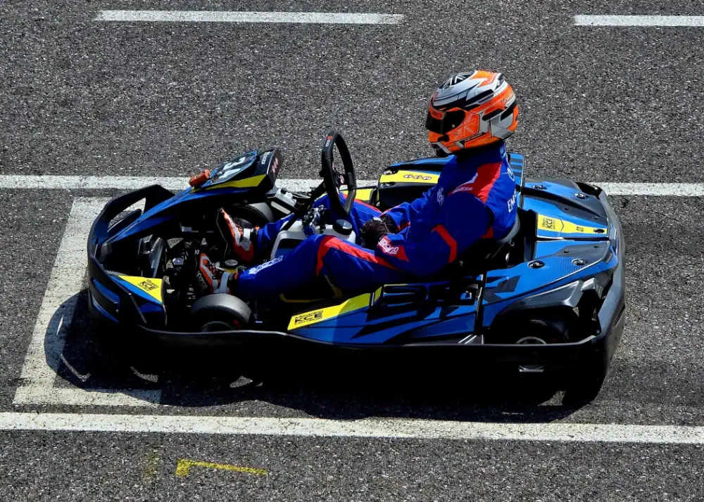 A person in a blue racing suit and orange helmet sits in a go-kart on an asphalt track, positioned between white starting lines.