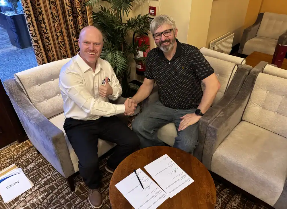 Two men are sitting in armchairs, shaking hands and smiling, with signed documents and pens on a round wooden table between them.