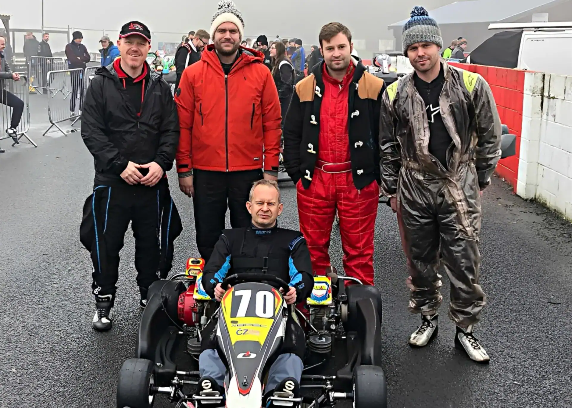 Five men pose together at an outdoor go-kart track; one is seated in a go-kart labelled 70 while the others stand behind him in racing suits and casual jackets.