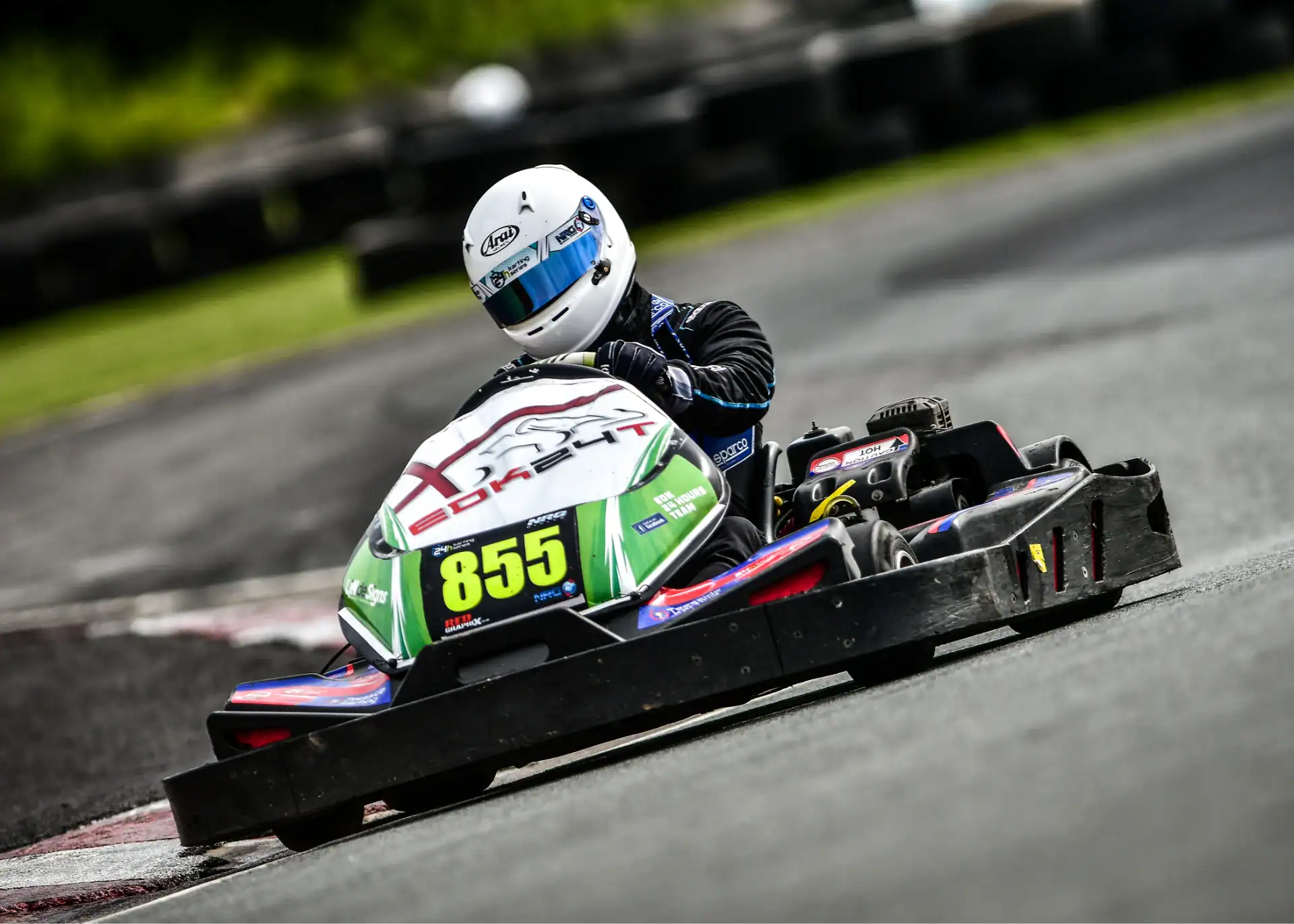A go-kart racer wearing a helmet drives kart number 855 on an outdoor track, leaning into a corner.