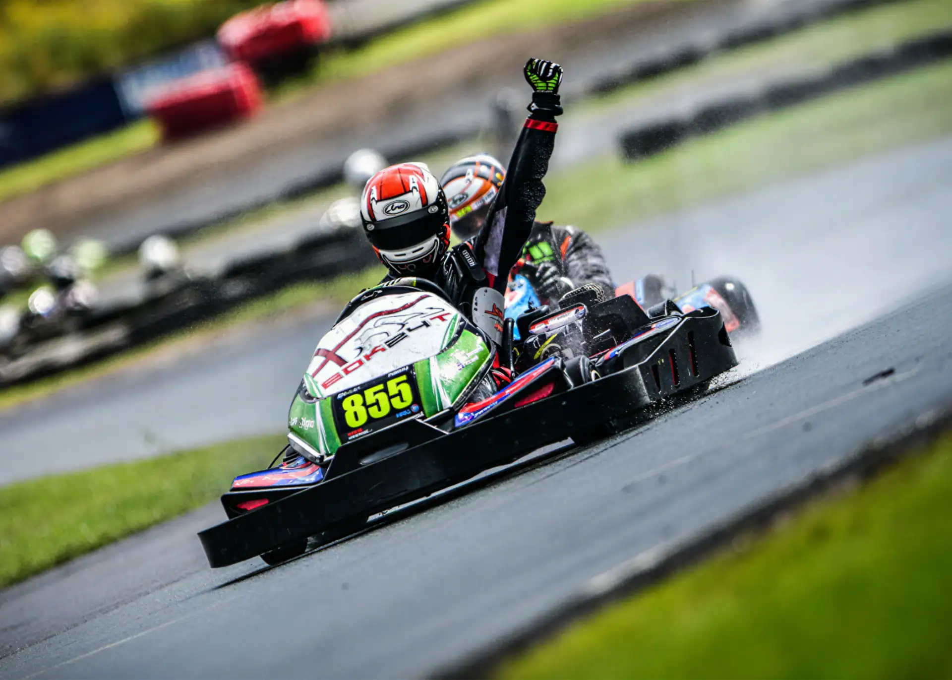 A go-kart driver wearing a helmet raises a fist in celebration whilst leading on a wet race track, with another driver close behind.