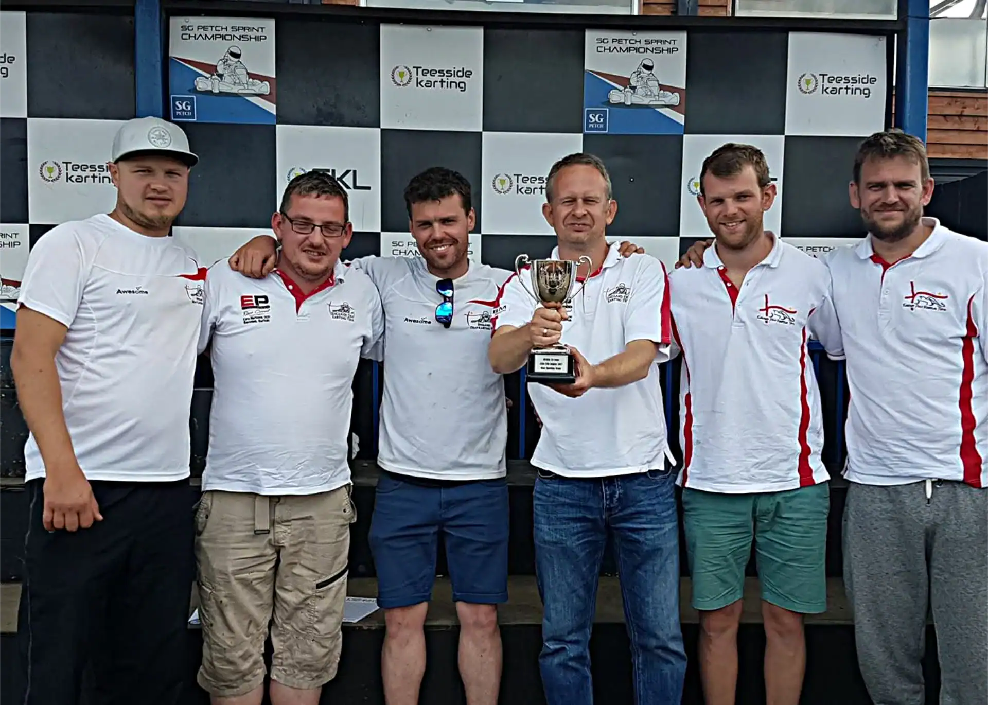 Six men in matching white polo shirts stand in front of a podium backdrop, with one man holding a trophy and the others posing alongside him.