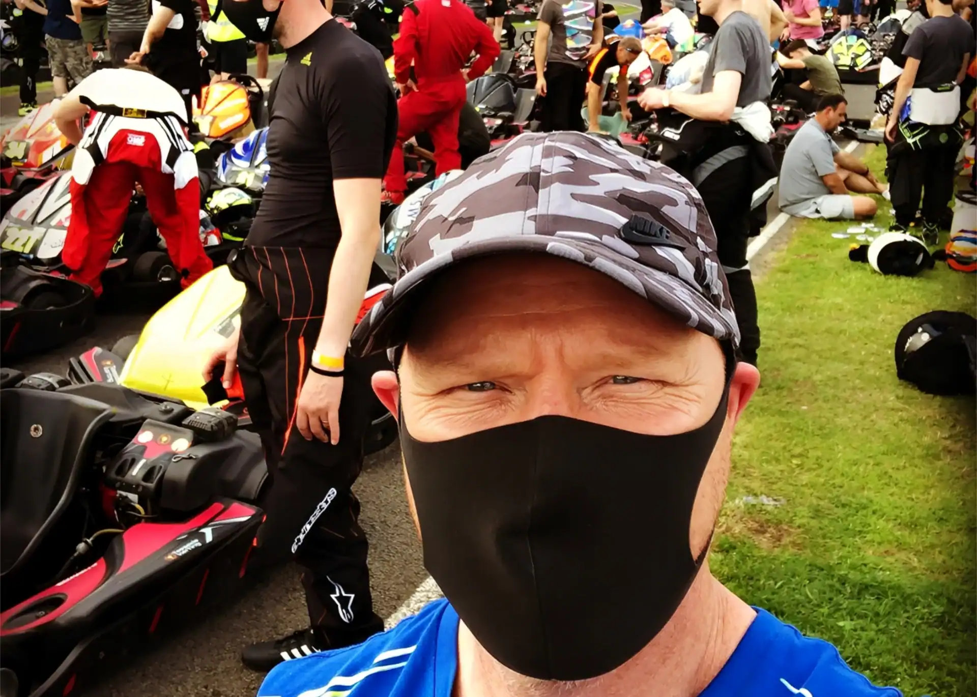 A man wearing a camo cap and black mask takes a selfie at an outdoor go-karting event with multiple people and karts in the background.
