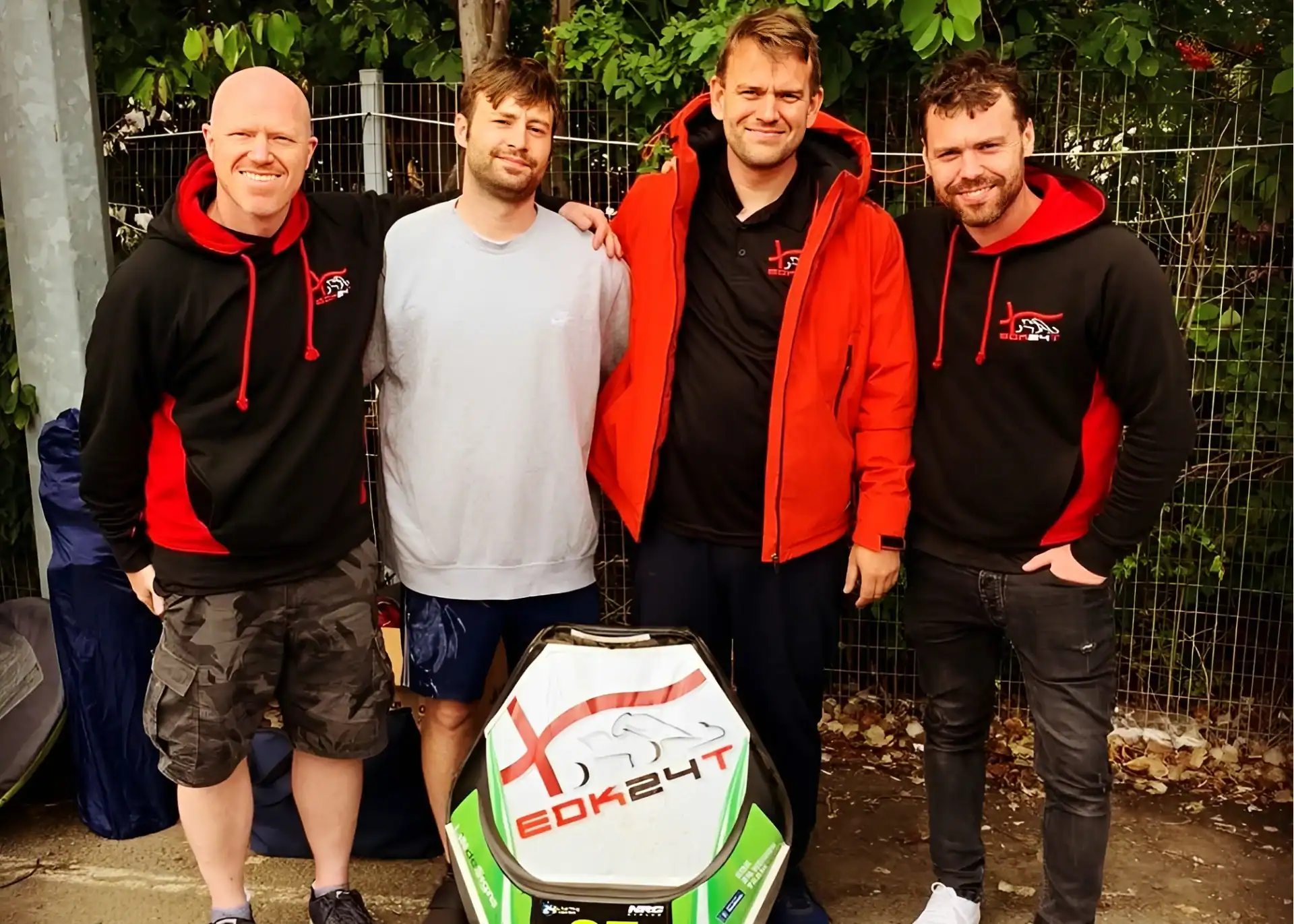 Four men stand outdoors beside a soapbox car labelled "EDK24T," with a fence and greenery in the background. Three are wearing matching black and red hoodies.