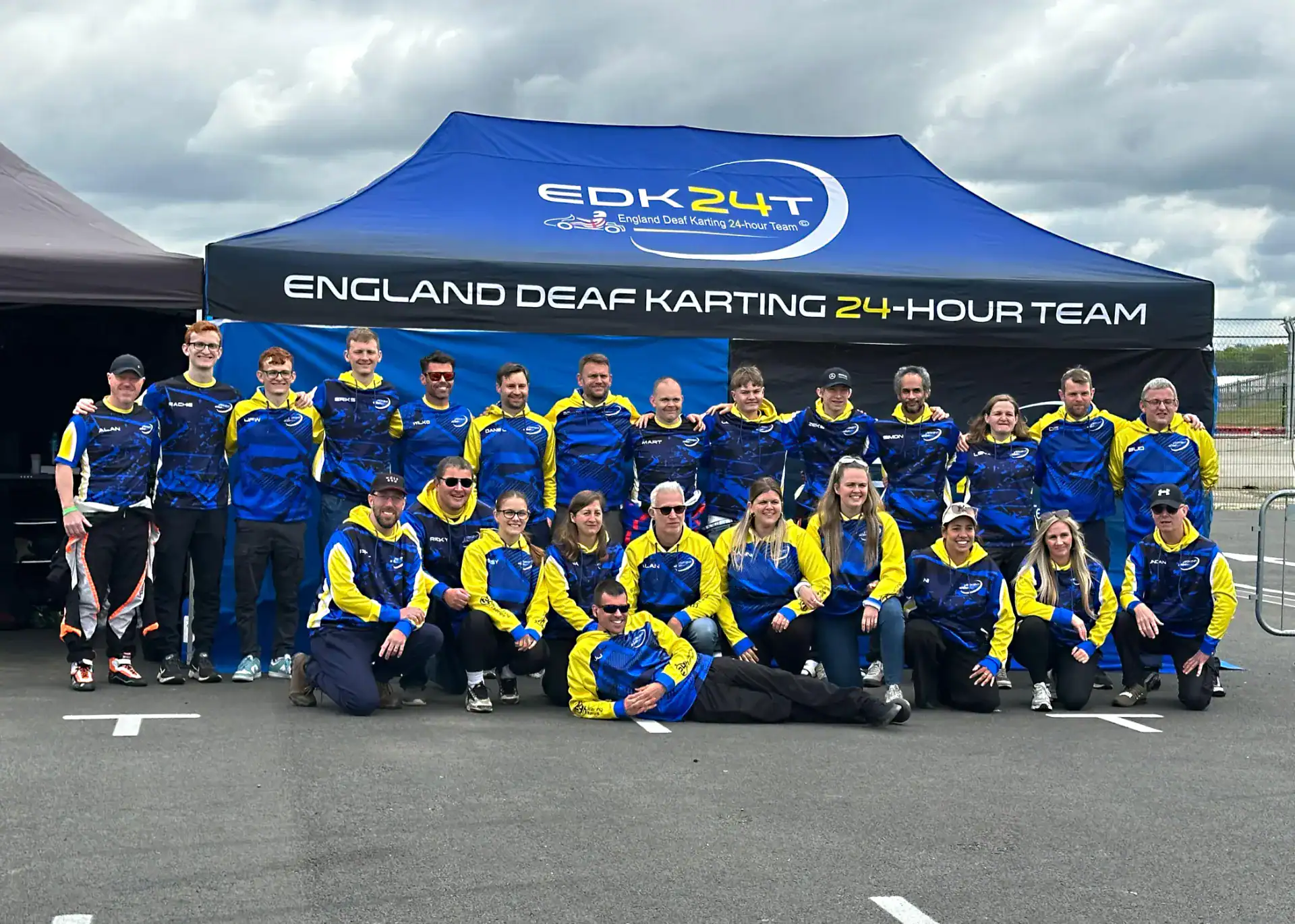 A group of people in matching blue and yellow racing team uniforms pose in front of a tent labelled "England Deaf Karting 24-Hour Team.