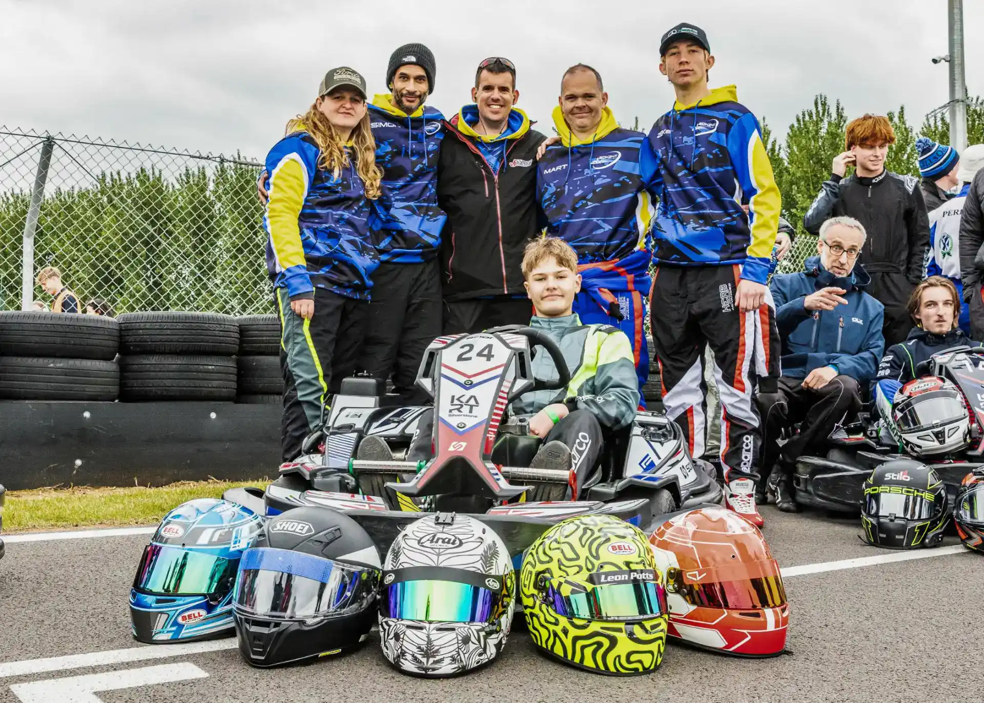 A group of people pose around a go-kart on a racetrack, with five racing helmets lined up in front of them and others standing or sitting nearby.