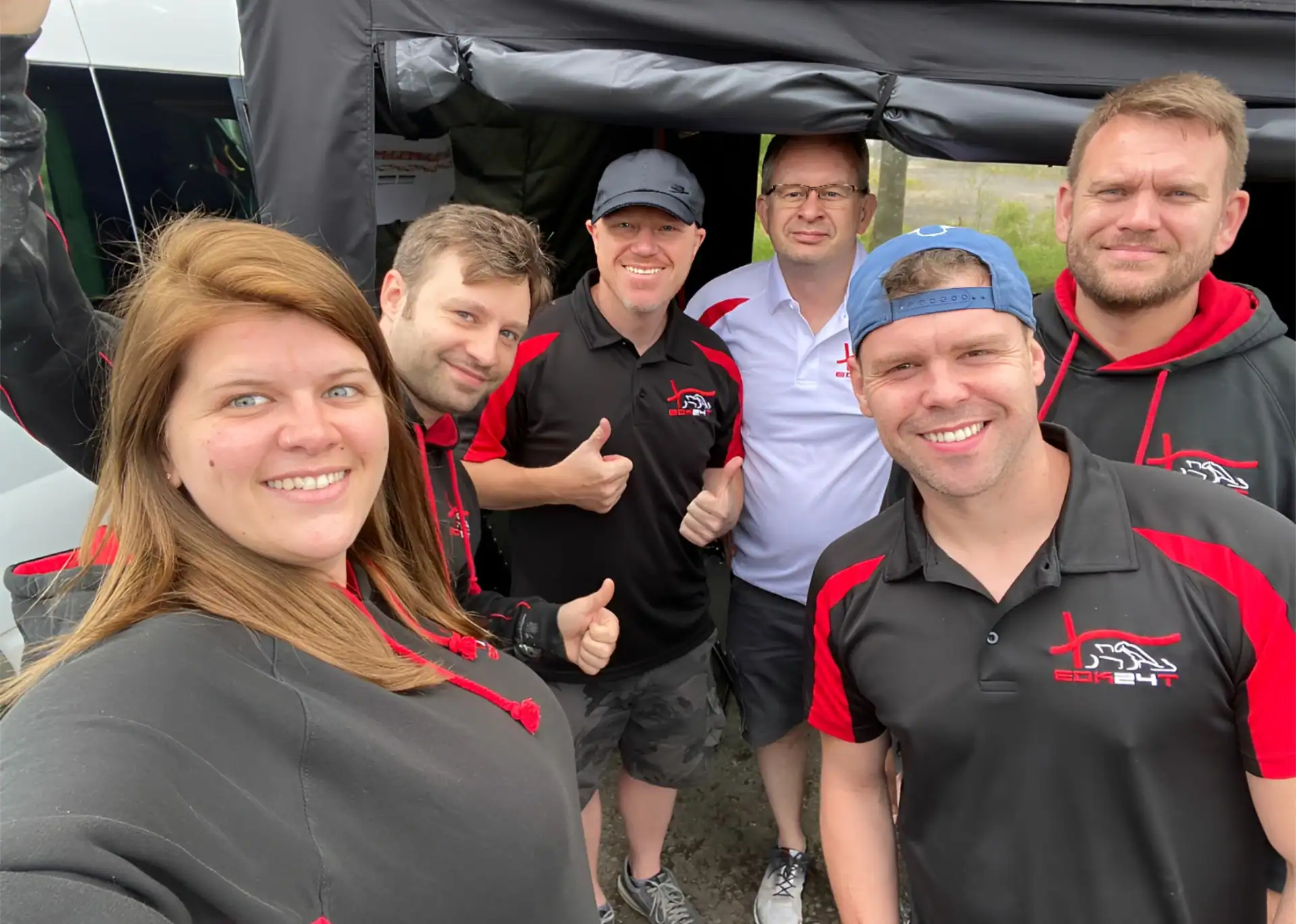 Six adults, five men and one woman, stand together outdoors smiling at the camera. Most wear black and red shirts with a logo, and one man wears a white shirt.