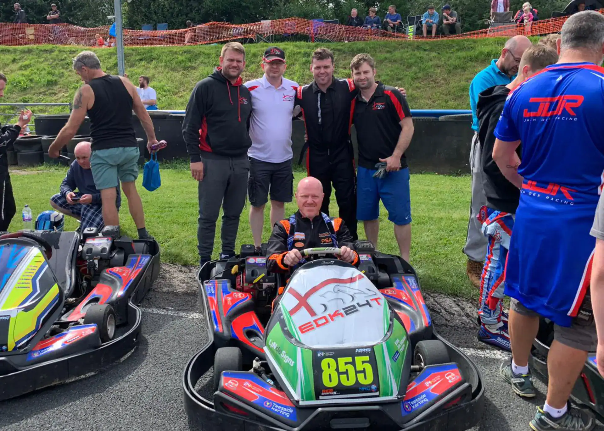 A man in a racing suit sits in a go-kart labelled 855, surrounded by a group of people standing on an outdoor track.