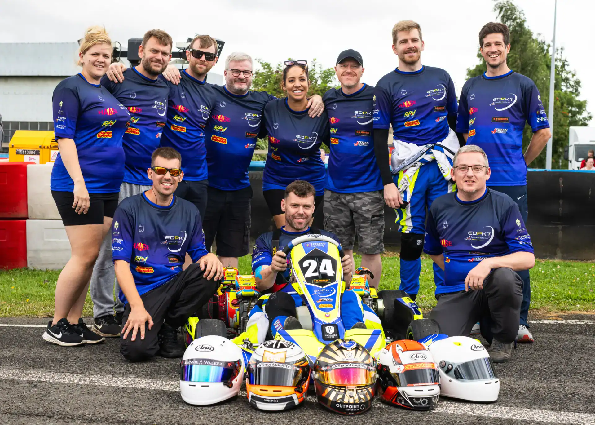 A group of twelve people, most wearing matching blue racing shirts, pose on a racetrack behind a go-kart with the number 24 and several racing helmets lined up in front.