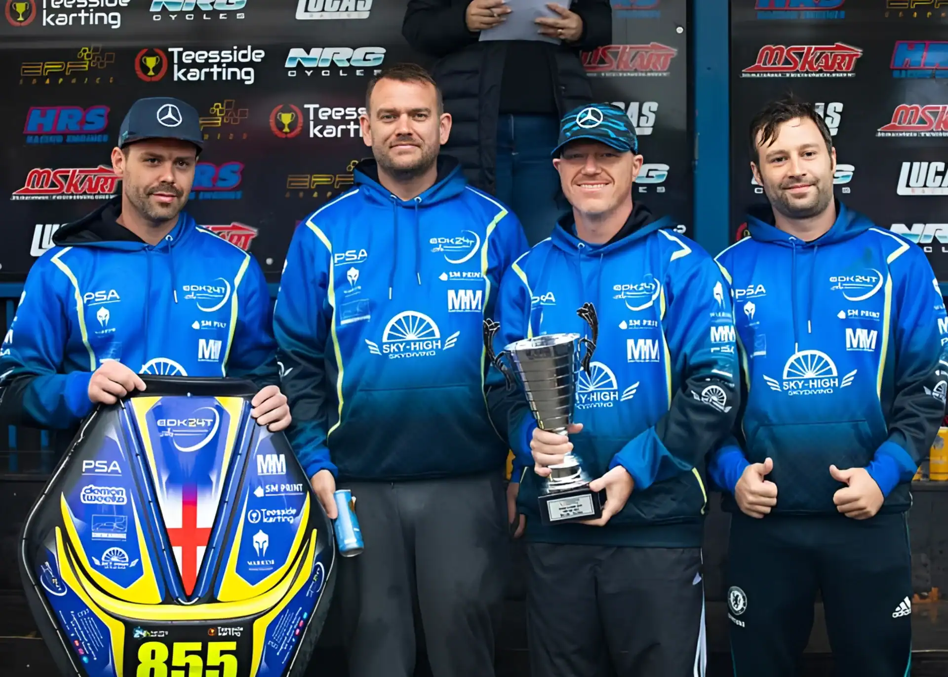 Four men in matching blue team jackets pose with a go-kart and a trophy at an outdoor racing event.