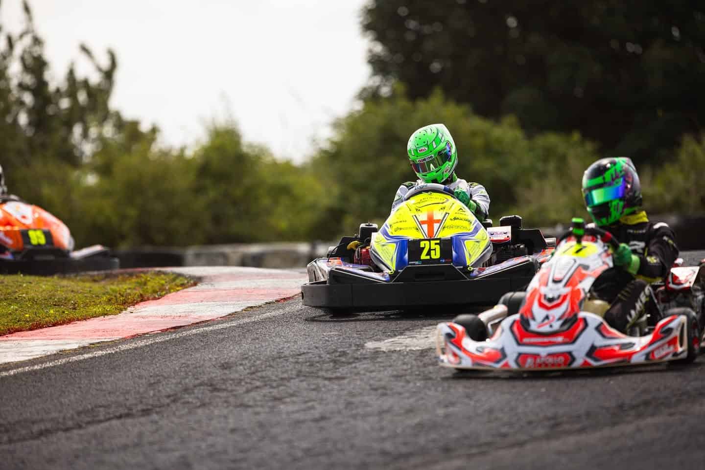 Three go-kart racers wearing helmets and racing suits navigate a curved track, with kart number 25 in the centre foreground. Trees and tyre barriers are visible in the background.