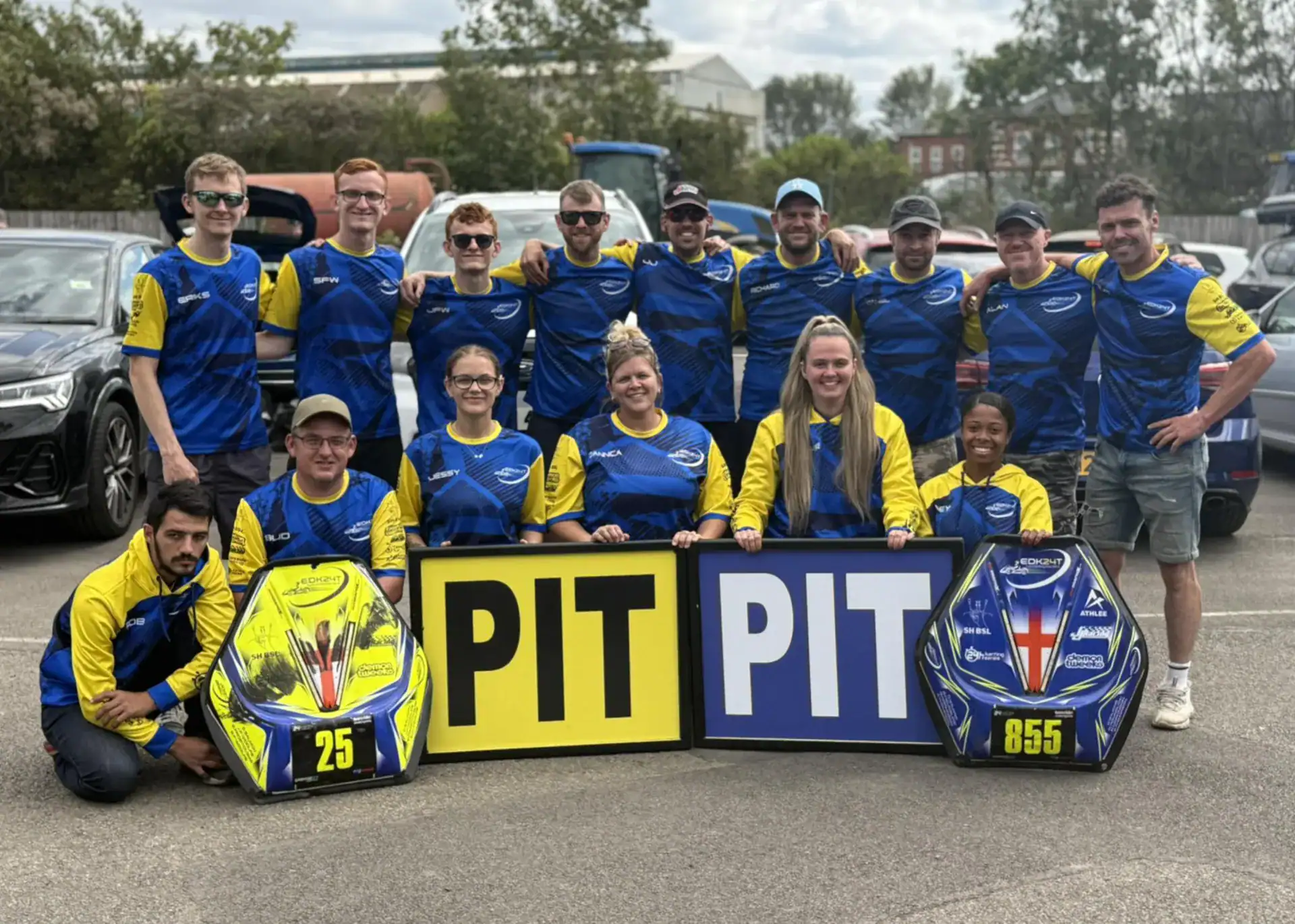 A group of 15 people in blue and yellow shirts pose outdoors behind two pit boards and two go-kart nose cones labelled 25 and 855.