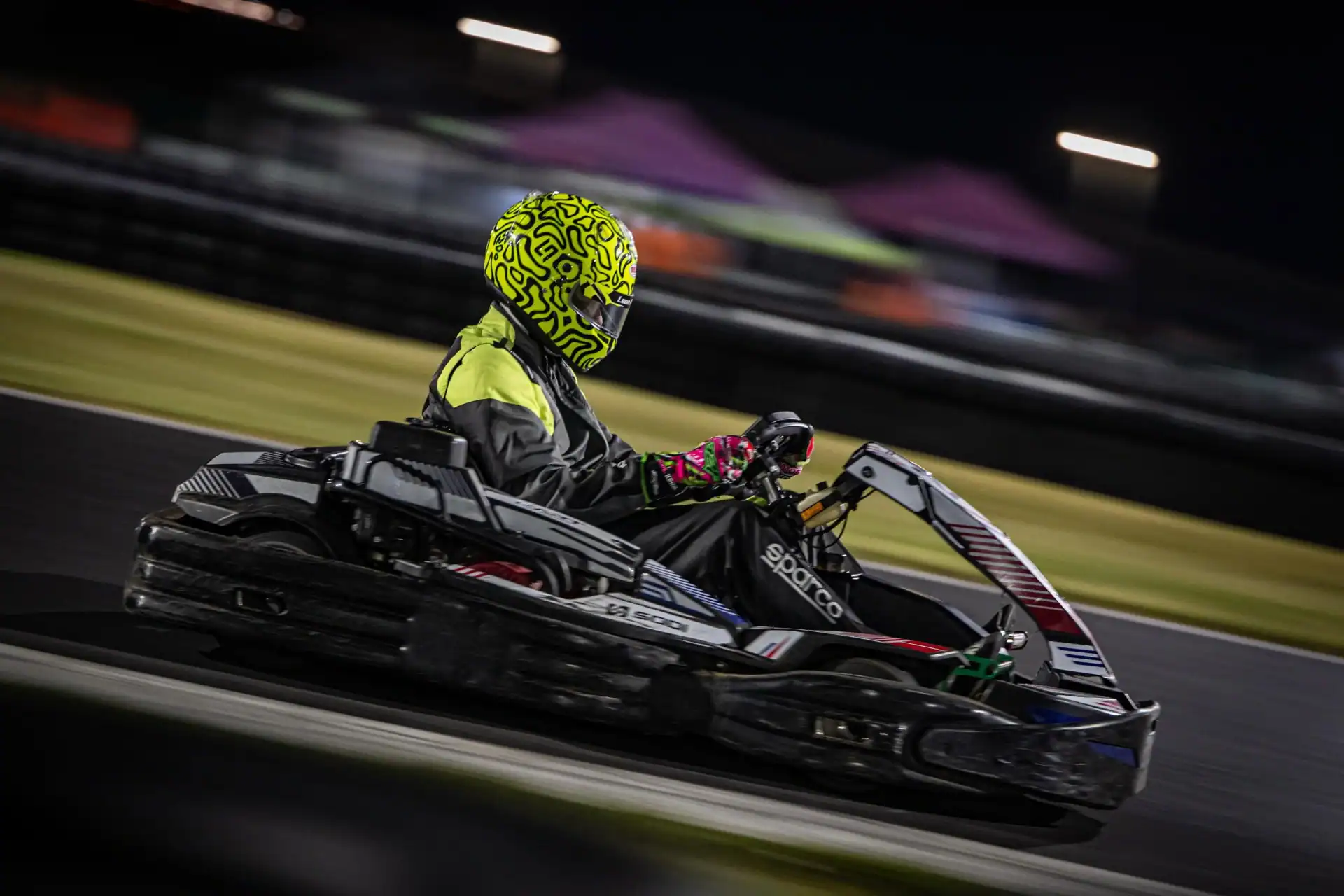 Person wearing a yellow patterned helmet and racing suit drives a go-kart on a track at night, background is blurred from motion.