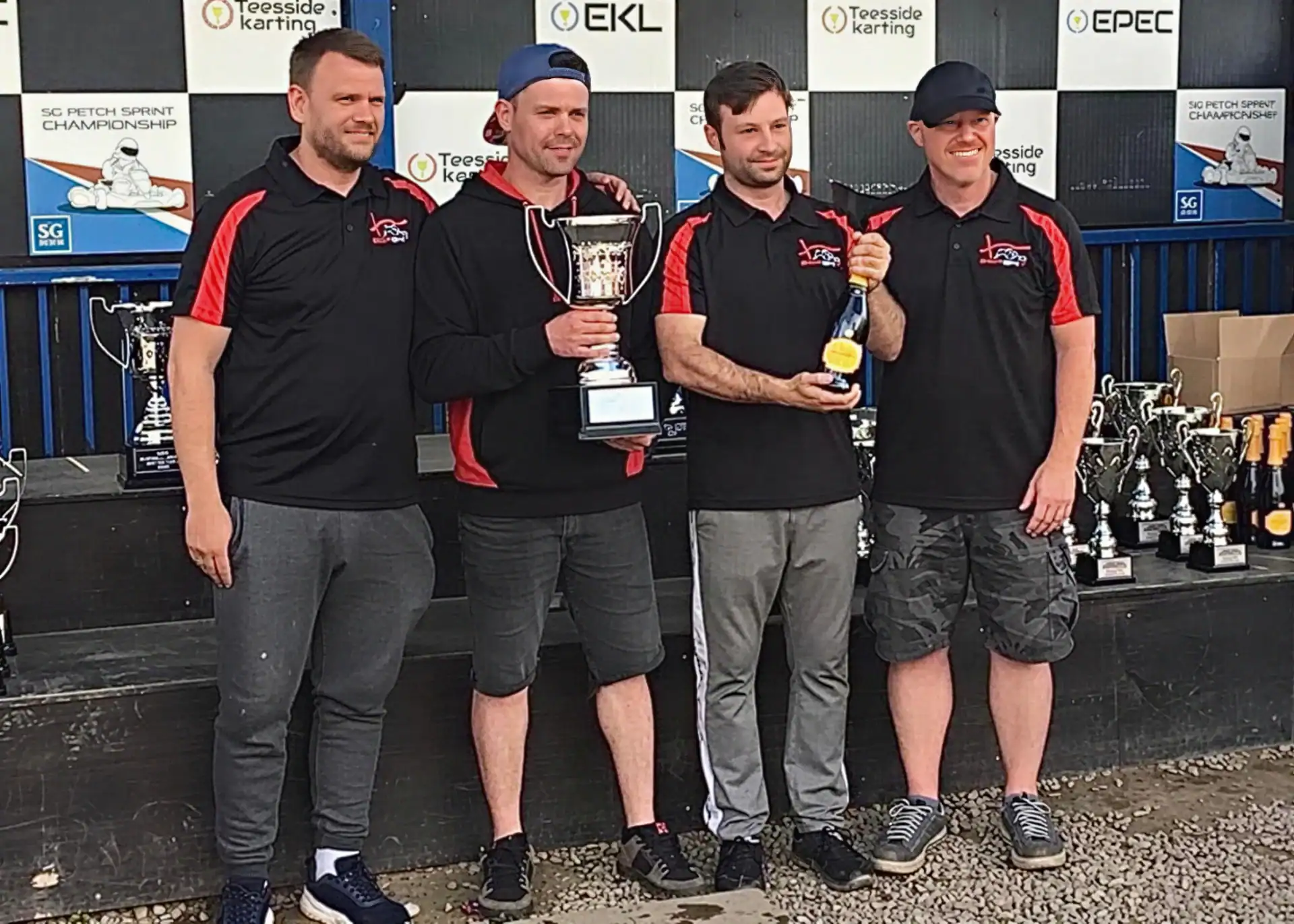 Four men in matching team shirts pose with trophies and medals on a podium at a karting championship event, with more trophies displayed on a table behind them.