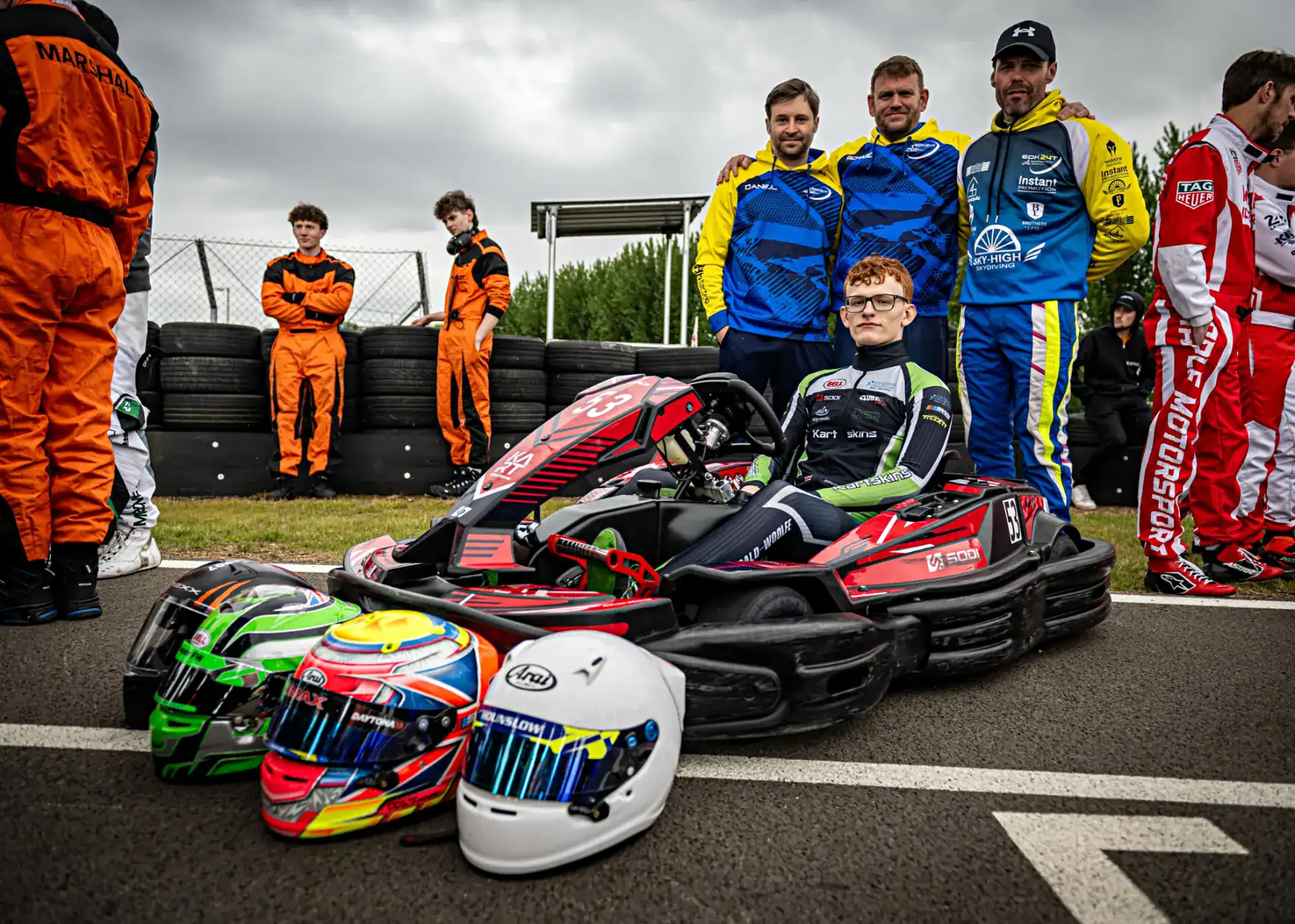 A young racer sits in a go-kart on a track with three helmets in front, surrounded by teams and officials standing in the background.