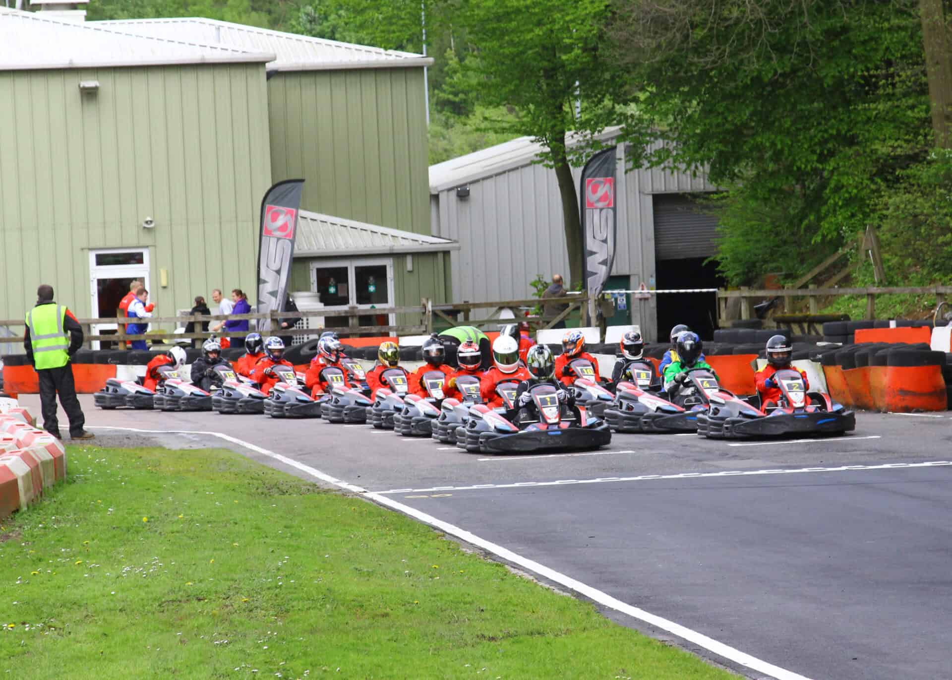 A group of go-kart racers in helmets and matching suits queue up at the starting line on an outdoor circuit, with a marshal standing nearby.