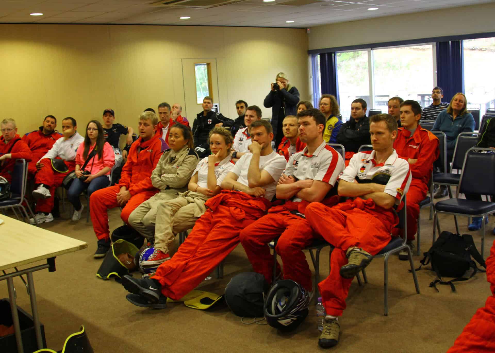A group of people in red and beige uniforms sit attentively in a room, facing a speaker or presentation at the front.