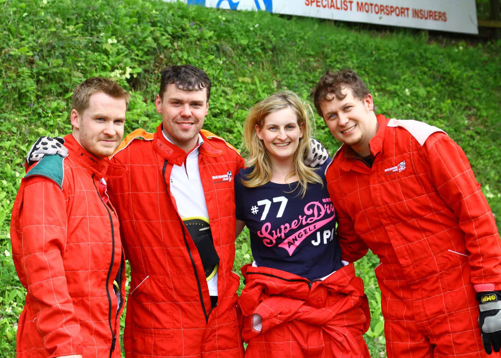 Four people, three wearing red racing suits, stand outdoors on grass smiling at the camera. A motorsport insurance sign is partially visible in the background.