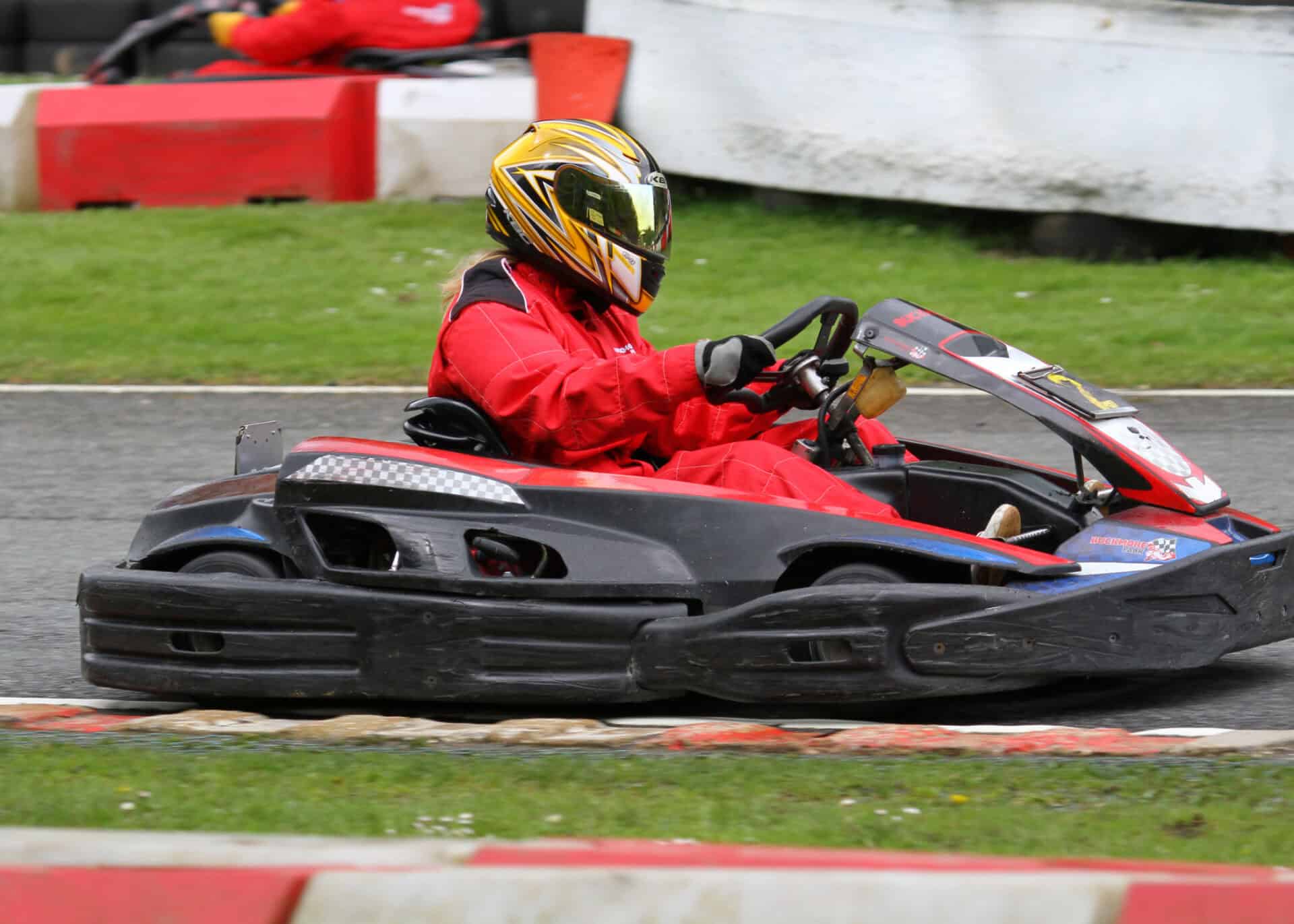 Person in a red racing suit and gold helmet driving a black go-kart on an outdoor racing track with grass and barriers in the background.