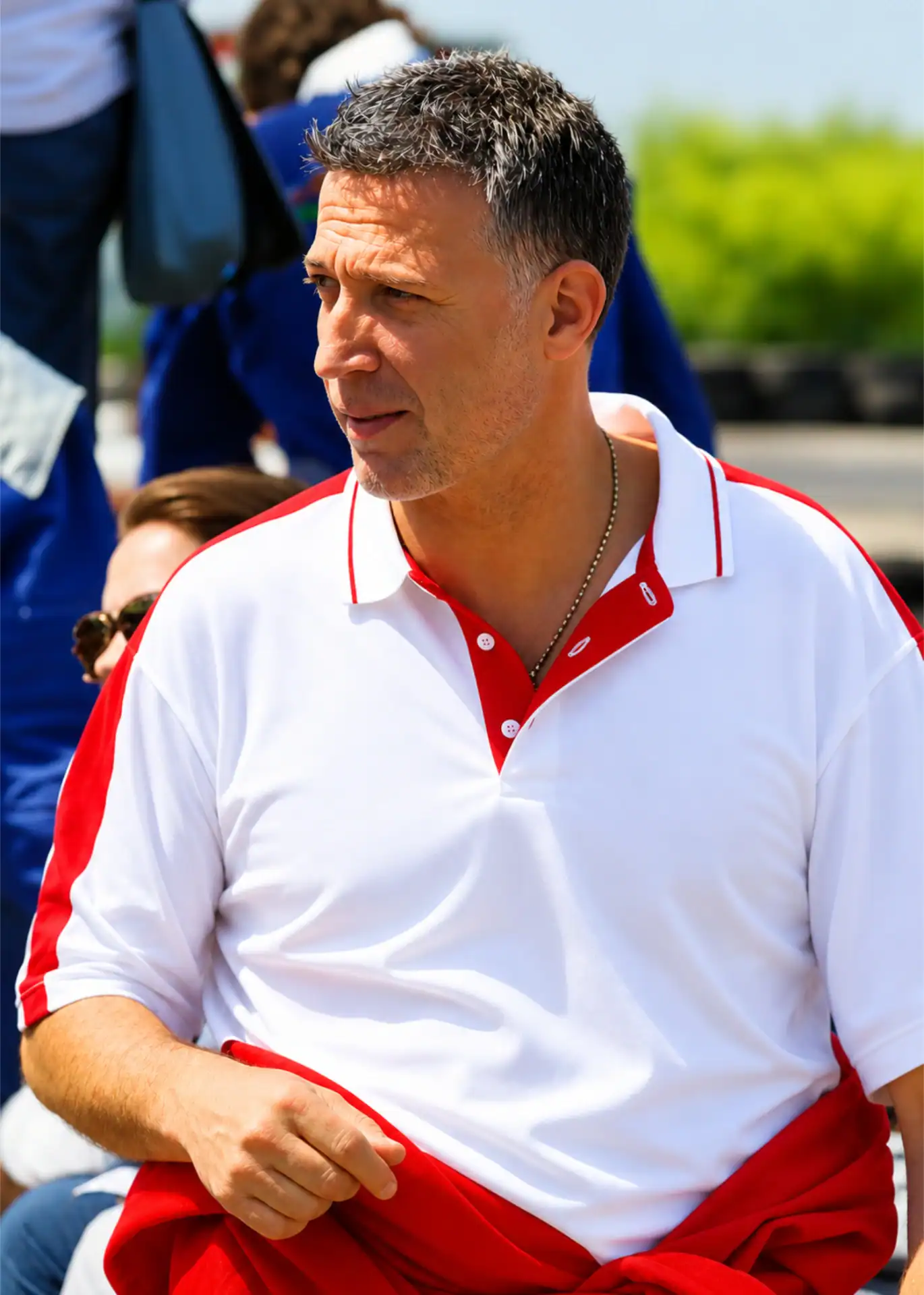 A man wearing a white polo shirt with red accents stands outdoors, holding a red jacket, with blurred people and greenery in the background.
