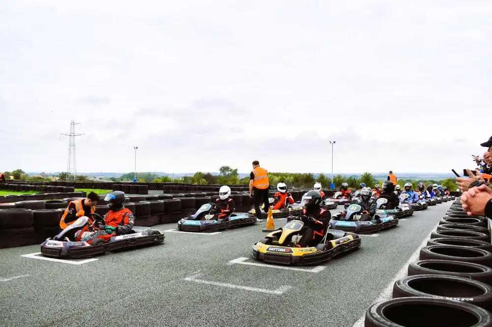 Go-kart drivers in helmets and gear line up on a starting grid, with officials in orange vests standing nearby and spectators on the right-hand side.