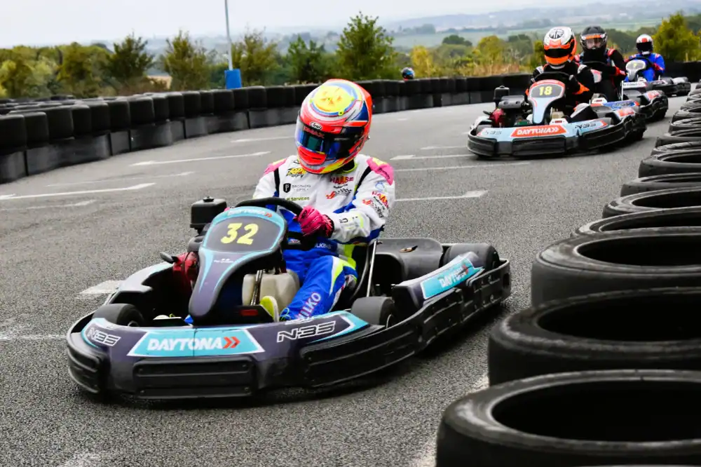 Go-kart racers wearing helmets and racing suits drive on an outdoor track bordered by stacked tyres, with the lead kart displaying the number 32.
