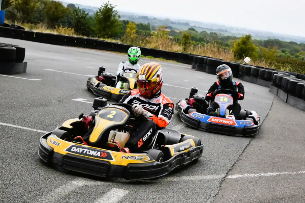 Three people wearing helmets race go-karts on an outdoor track, with tyres lining the edge and trees visible in the background.