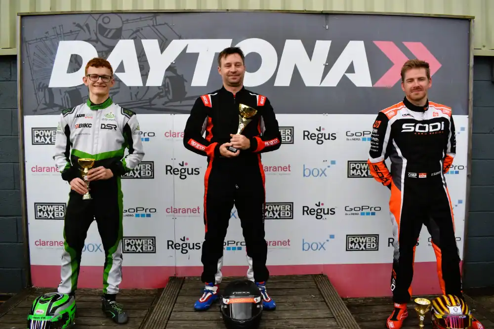 Three go-kart racers stand on a winners’ podium holding trophies at a Daytona event, with sponsor logos displayed on the backdrop behind them.